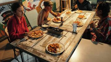 A group of people dining at a rustic wooden table with various dishes and a fan in the background.