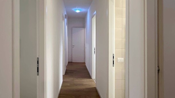 Ein schmaler Flur mit weißen Wänden und Holzboden, der zu einer verschlossenen Tür am Ende führt.