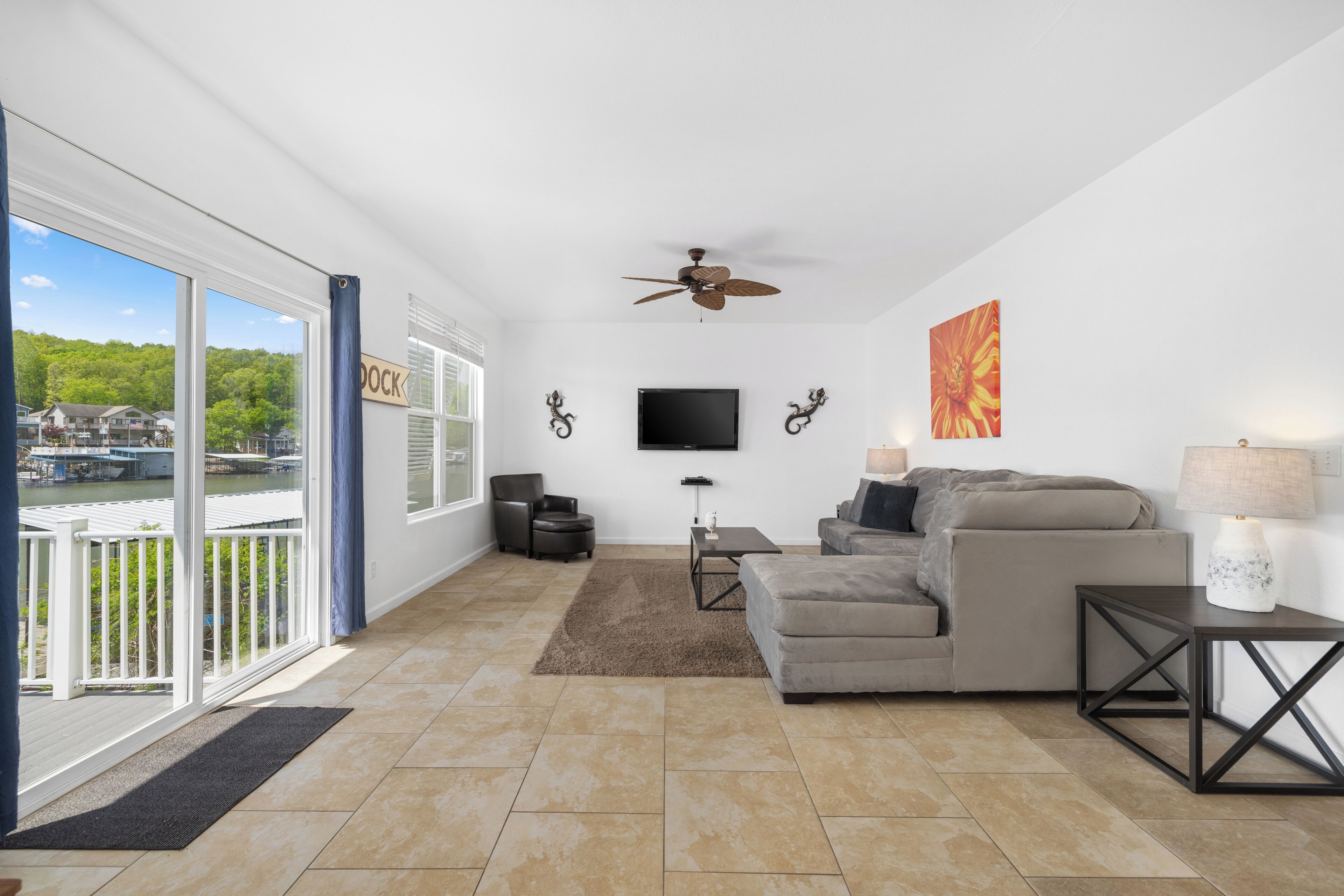 A spacious living room with a large window, a ceiling fan, a flat-screen TV, a gray sofa, a coffee table, and a side table with a lamp.