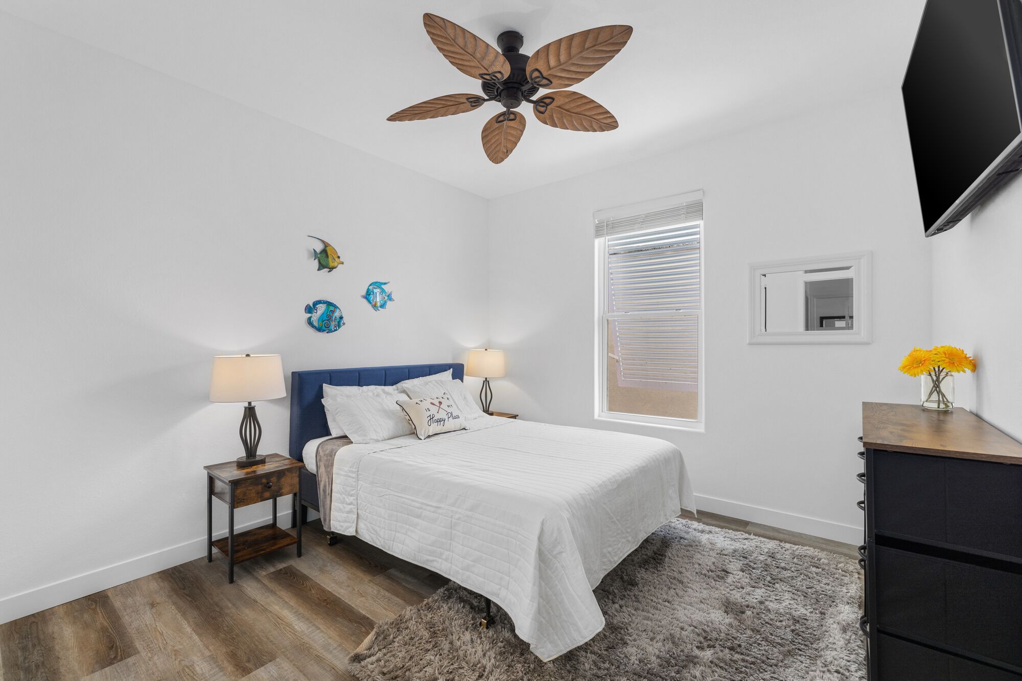 A bedroom with a bed, two bedside tables, a ceiling fan, a window with blinds, decorative fish wall art, and a TV mounted on the wall.