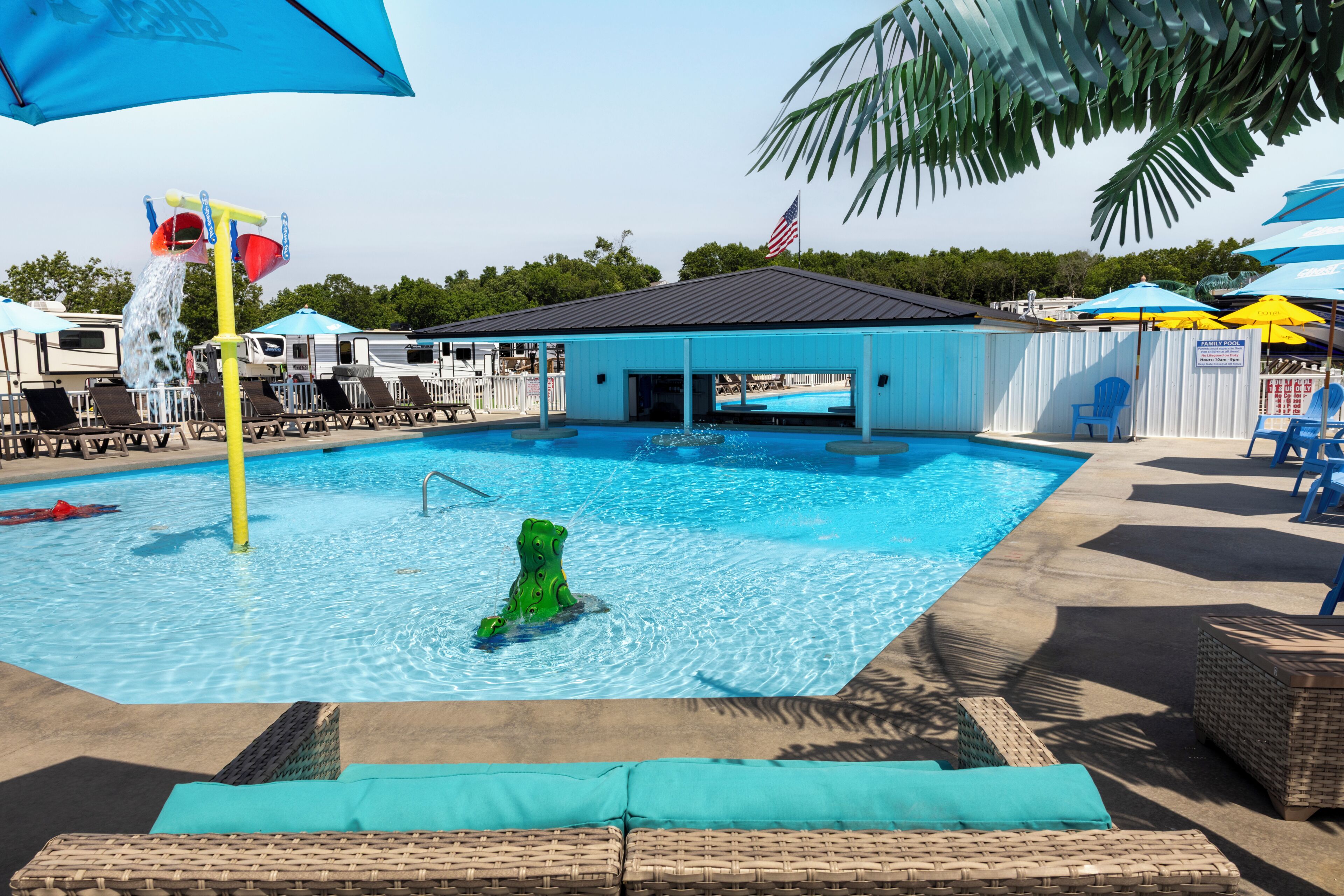 Una piscina con zona de juegos acuáticos, sillas de descanso y un pabellón cubierto.