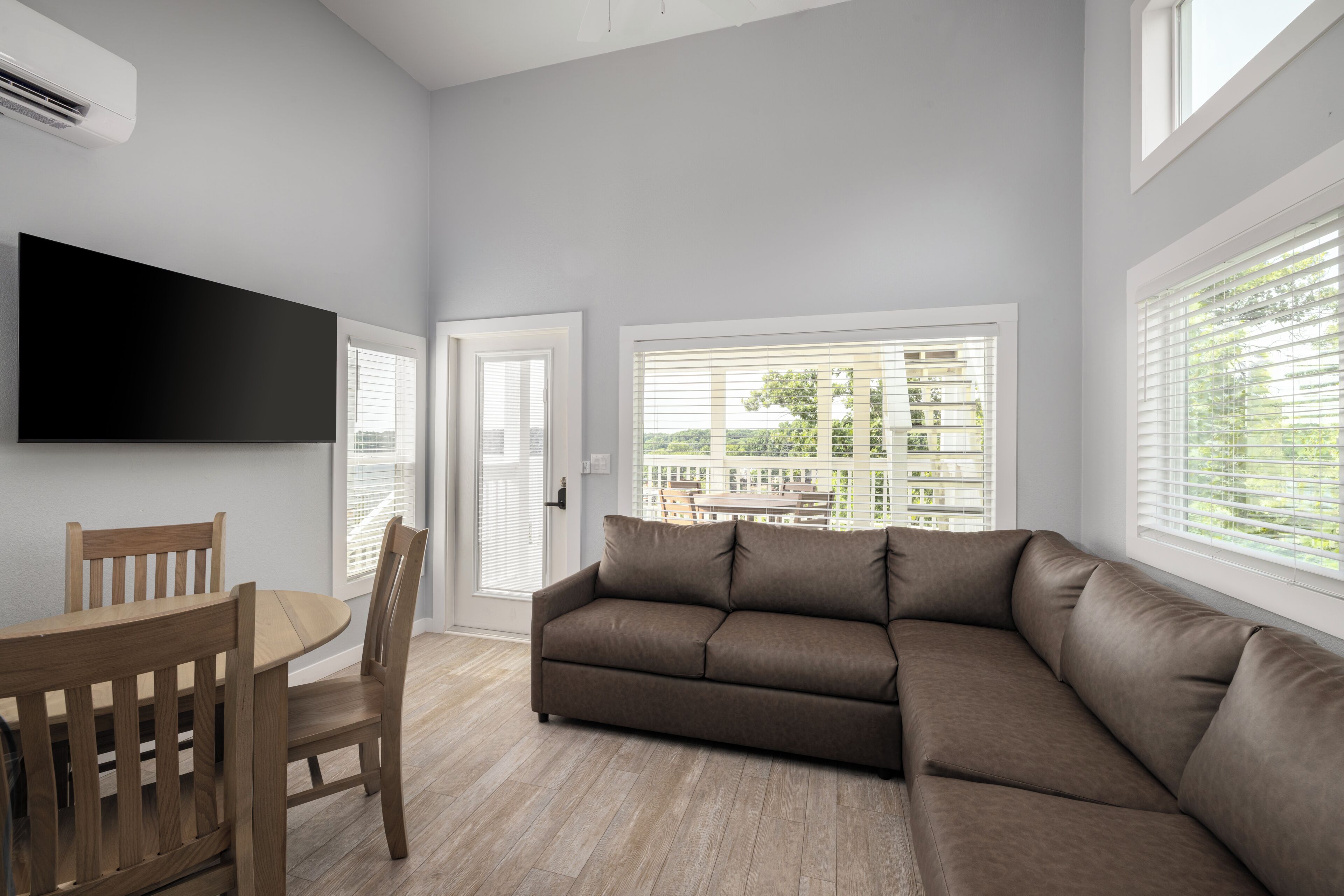 A modern living room with a sectional sofa, a dining table with chairs, a flat-screen TV mounted on the wall, and large windows with blinds.
