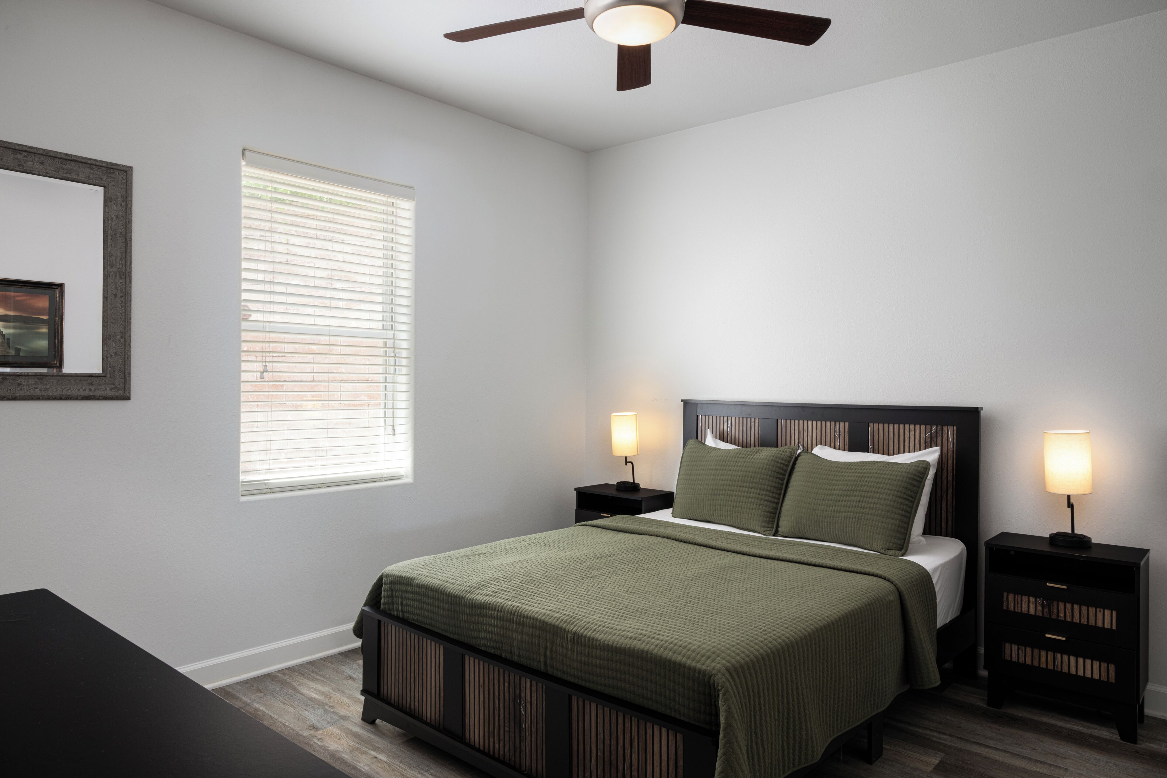 A bedroom with a bed, two bedside tables with lamps, a ceiling fan, a window with blinds, and a framed picture on the wall.