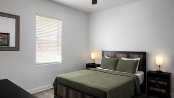 A bedroom with a bed, two bedside tables with lamps, a ceiling fan, a window with blinds, and a framed picture on the wall.