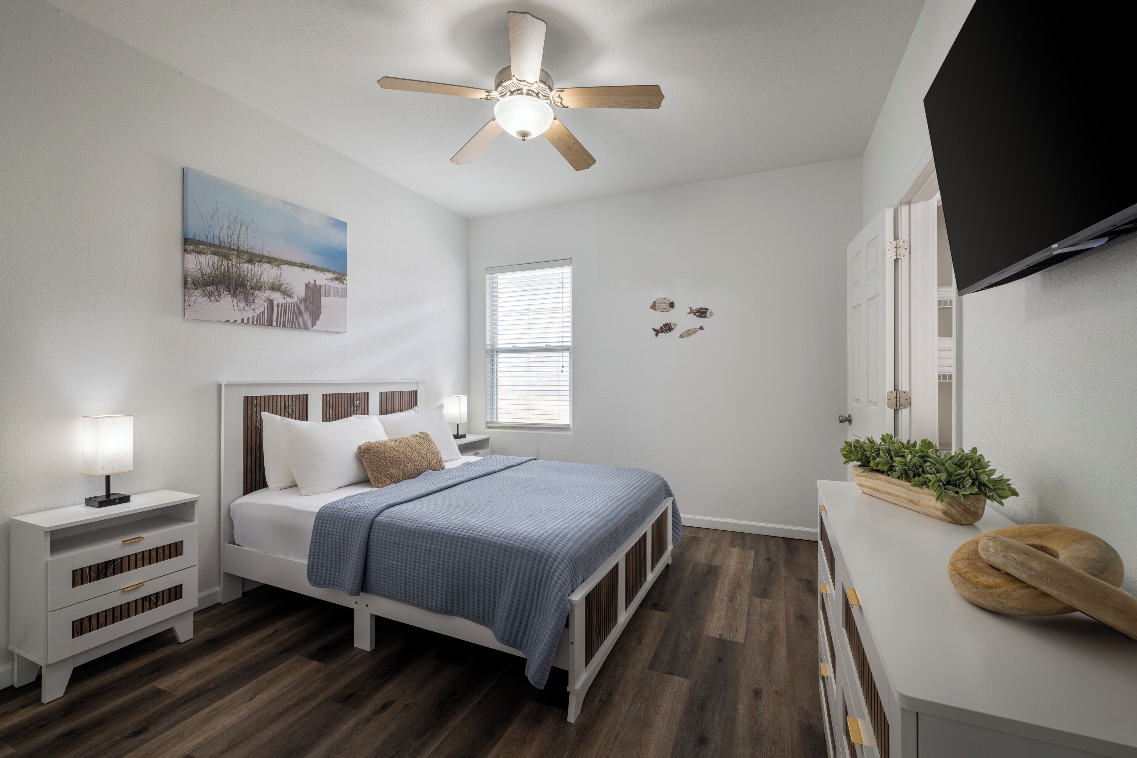 A bedroom with a bed, nightstands, a ceiling fan, a window, wall art, and a TV.