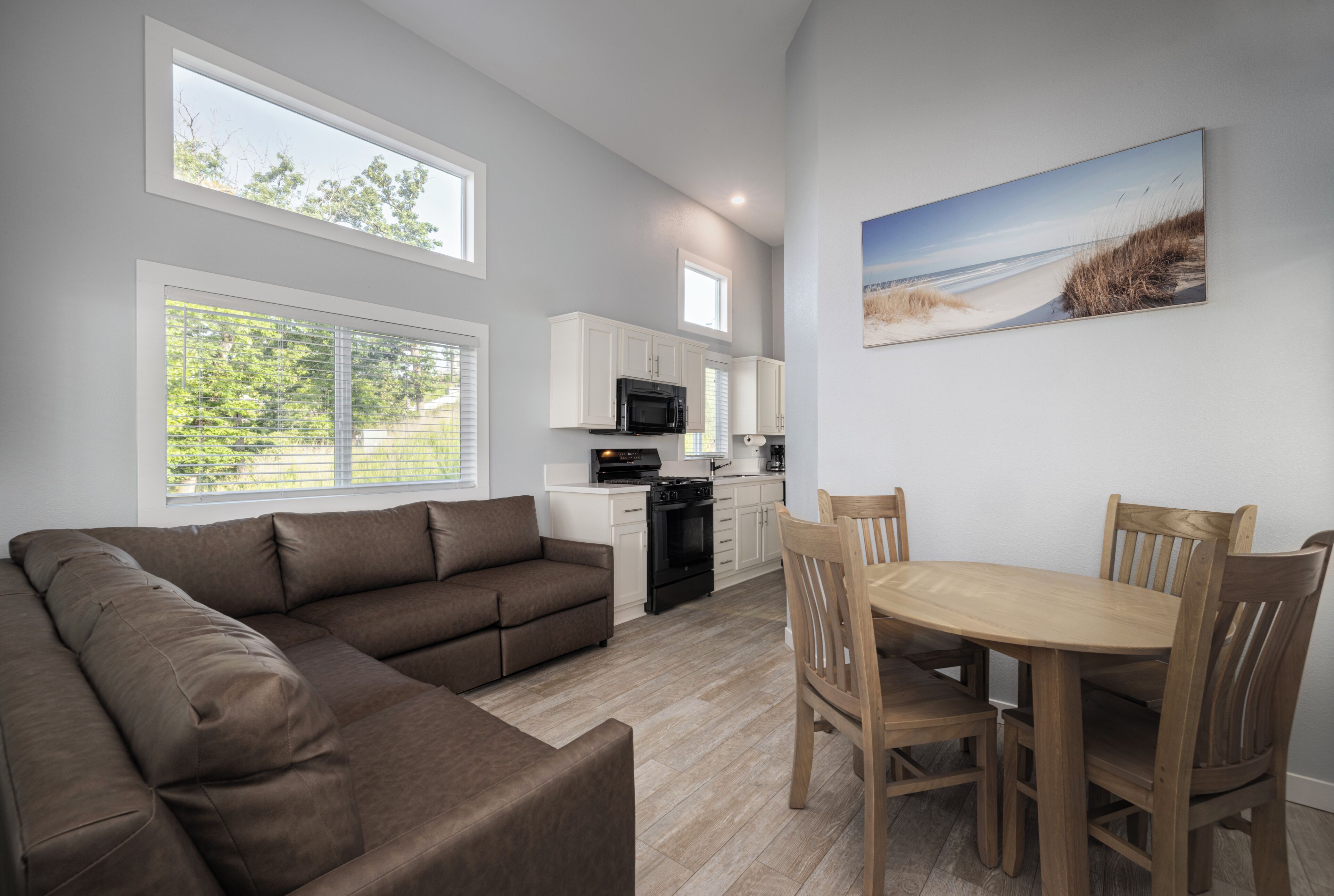 A modern kitchen with white cabinets, a dining table with chairs, a brown sofa, and large windows.