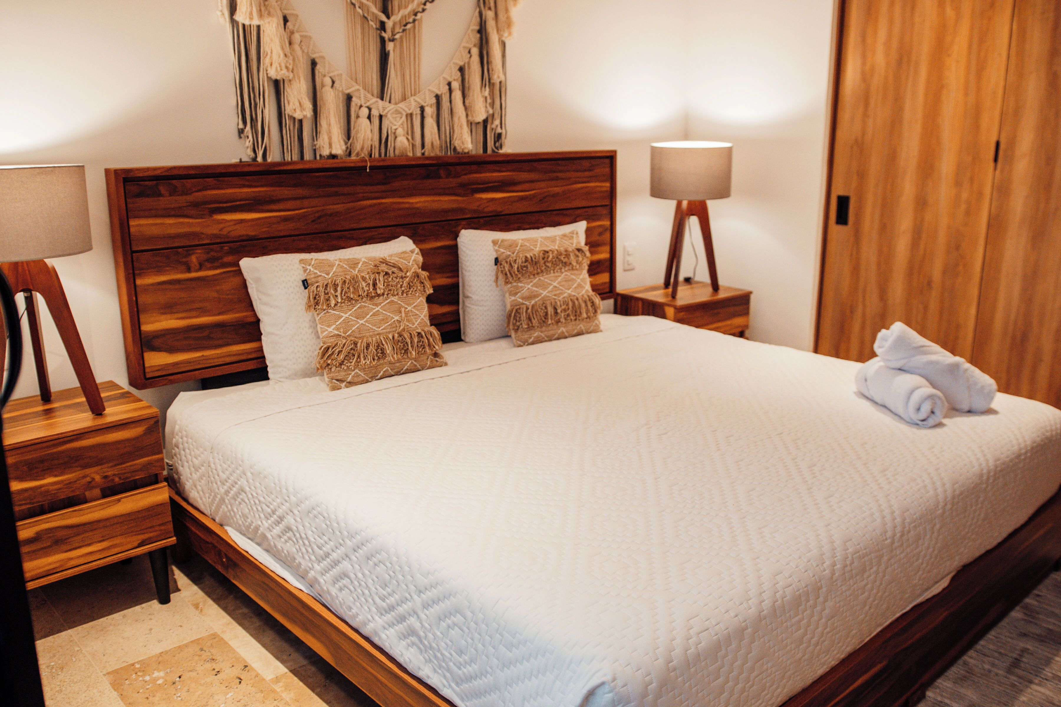 Een opgemaakt bed met een houten hoofdbord, twee nachtkastjes met lampen, en een wanddecoratie erboven.