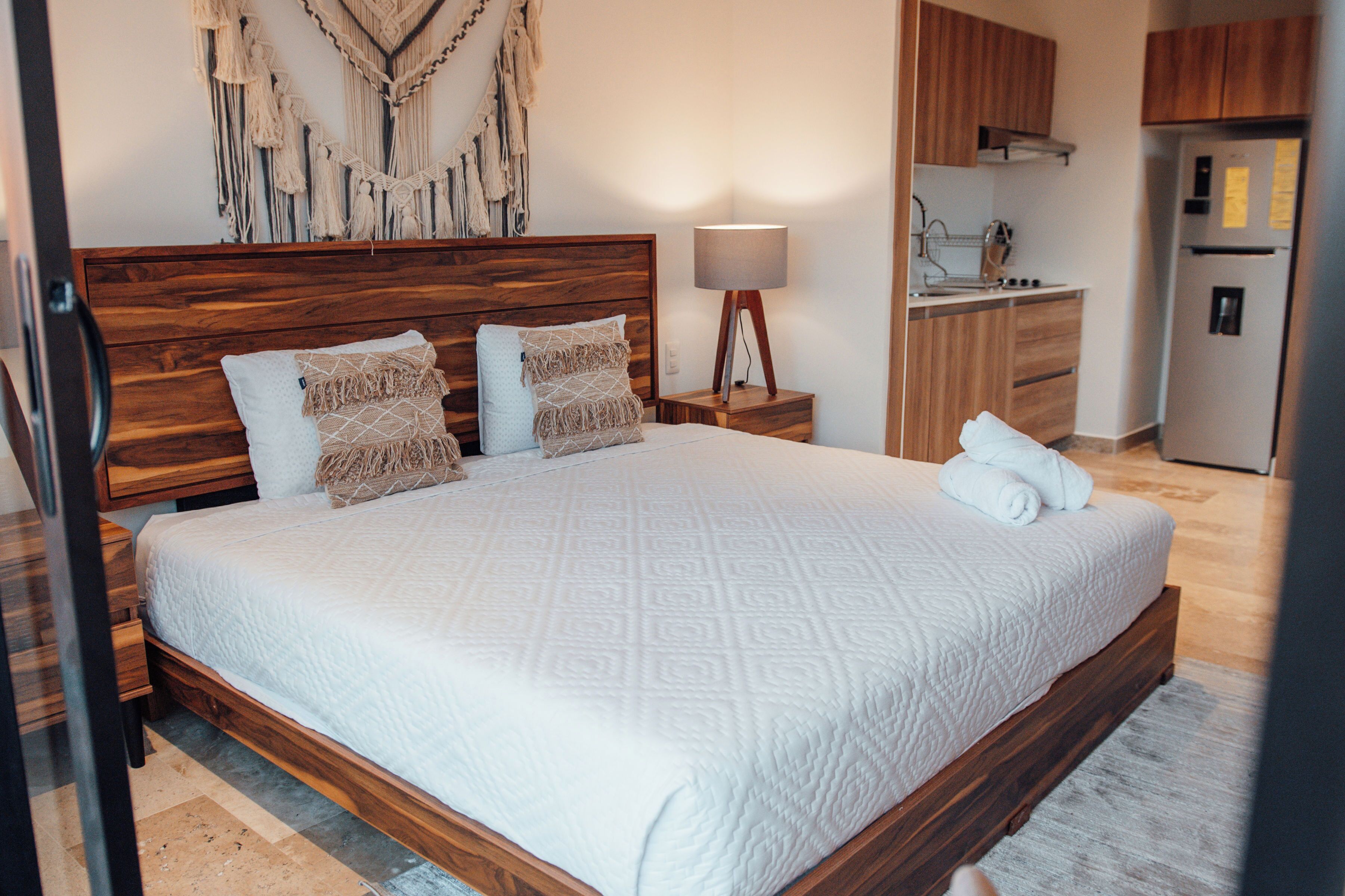 Une chambre à coucher avec un lit en bois, une table de chevet avec une lampe, et un coin cuisine visible à travers une porte ouverte.