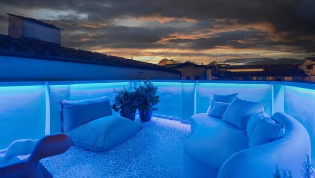Eine Dachterrasse mit einem blau beleuchteten Pool, weißen Gartenmöbeln und Blick auf die umliegenden Gebäude bei Sonnenuntergang.