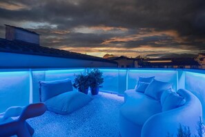 Une terrasse sur le toit avec une piscine illuminée de lumière bleue, des meubles d’extérieur blancs et une vue sur les bâtiments environnants au coucher du soleil.