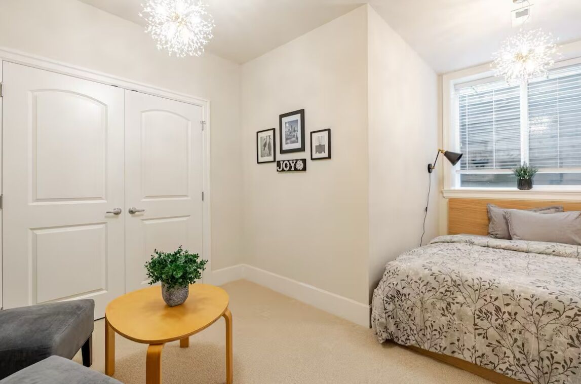 Une chambre avec un lit, une petite table sur laquelle est posée une plante, un fauteuil de bureau, une fenêtre avec des stores et un mur orné de tableaux et d’une enseigne portant le mot « JOIE ».