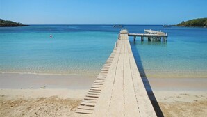 Une jetée en bois s’étendant dans des eaux calmes et d’un bleu limpide, avec une plage de sable à son extrémité.