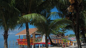 Un hôtel situé en bord de mer, doté d’une terrasse rouge et jaune, d’une jetée en bois et de palmiers.