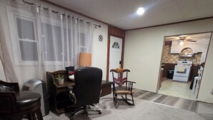 Ein Zimmer mit Teppichboden, einem Schreibtisch mit Stuhl, einem Schaukelstuhl, einem kleinen Tisch mit Lampe und einer Küchenzeile mit Herd und Backofen.