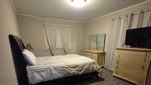 Ein Schlafzimmer mit Bett, Kommode, Fernseher und einem Fenster mit Vorhängen.
