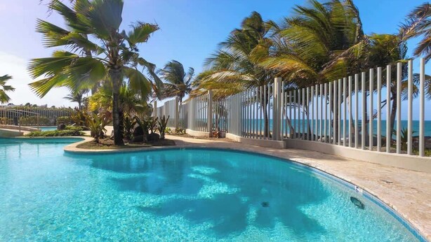 A clear blue pool with palm trees and a fence, surrounded by a clear sky. - Beachfront by Otium | Pool | BBQ | Beach Access (Río Grande)