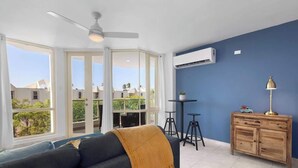 A modern living room with a sofa, a coffee table, and a wooden cabinet. There are large windows and an air conditioning unit on the wall. - Beachfront by Otium | Pool | BBQ | Beach Access (Río Grande)