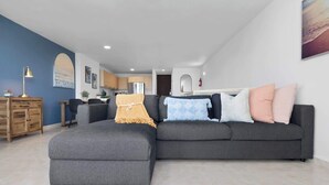 A modern living room with a grey sectional sofa, a wooden cabinet, and a kitchen in the background. - Beachfront by Otium | Pool | BBQ | Beach Access (Río Grande)