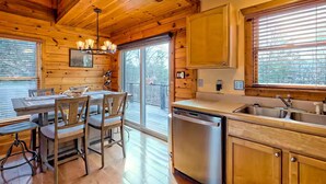 A wooden cabin interior with a dining table, chairs, a kitchen with a sink and dishwasher, and large windows.