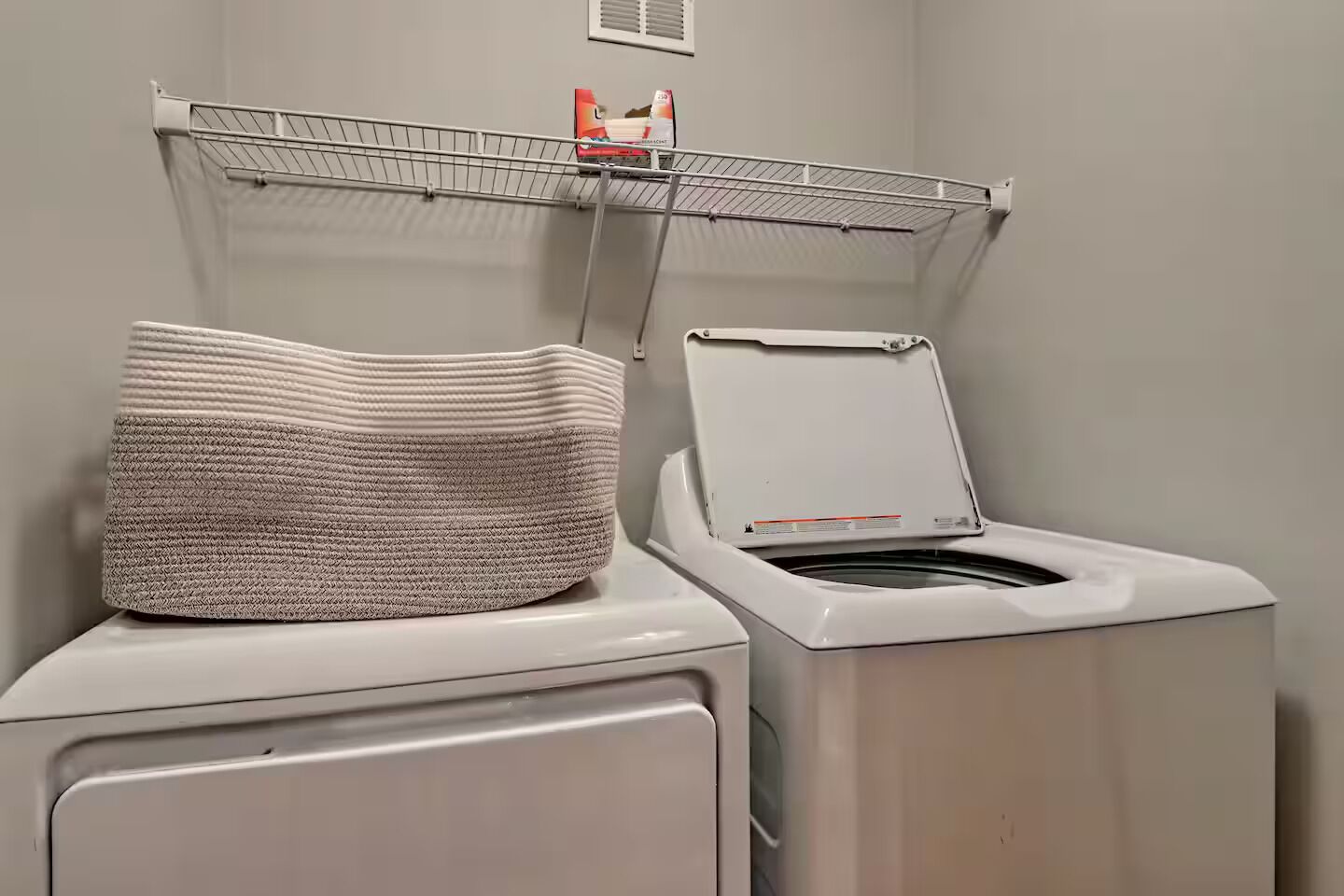 Un área de lavandería con lavadora y secadora, un estante de metal sobre la secadora y una cesta a rayas encima de la secadora.