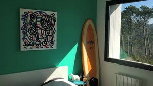 Une chambre avec un lit, une planche de surf appuyée contre le mur et une fenêtre donnant sur la forêt.