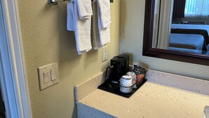 Une salle de bain avec un comptoir de couleur beige, une machine à café, un paquet de café et deux serviettes suspendues à un porte-serviettes.
