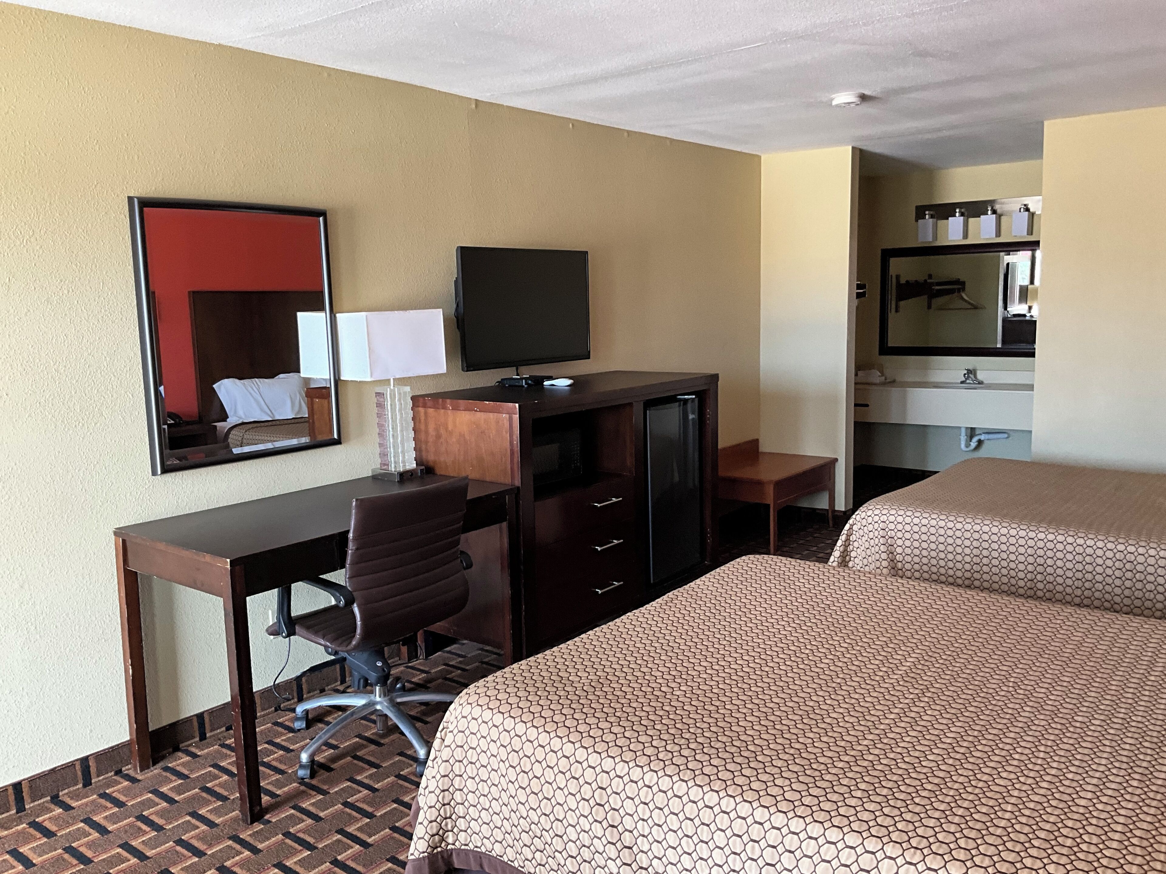 Ein Hotelzimmer mit Bett, Schreibtisch mit Stuhl, Fernseher, Spiegel und einem Badezimmer mit Waschbecken und Spiegel.