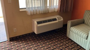Une pièce avec un tapis à motifs, un radiateur portatif, une fenêtre avec des rideaux légers et un fauteuil à motifs.