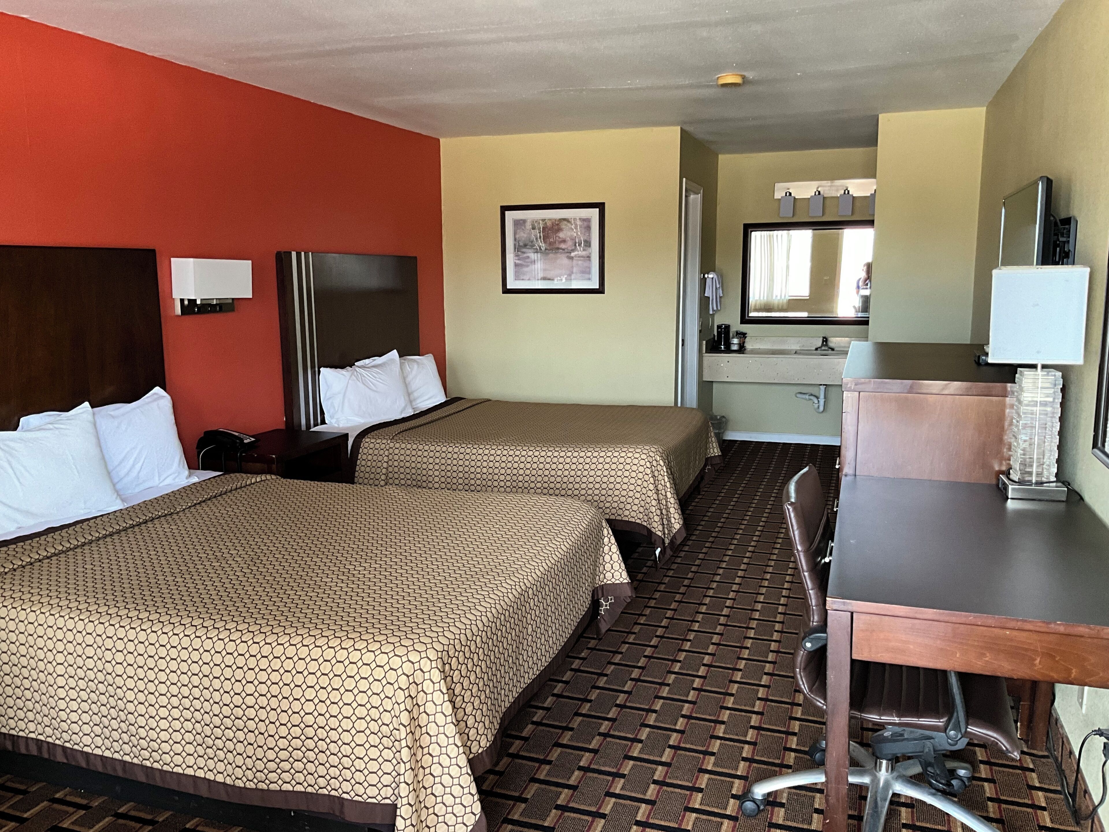 A hotel room with two beds, a desk, a chair, a sink, and a mirror.