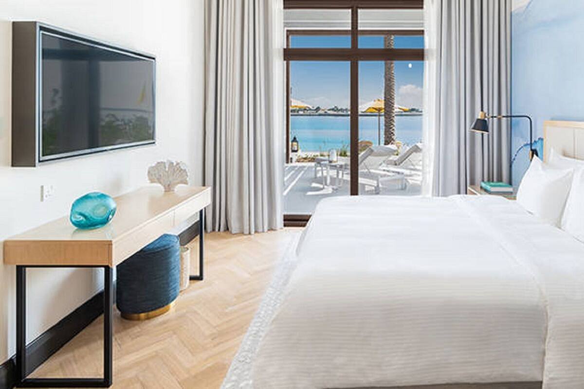 Habitación de hotel con cama, escritorio, televisor, vista al mar y balcón con sillones de descanso.
