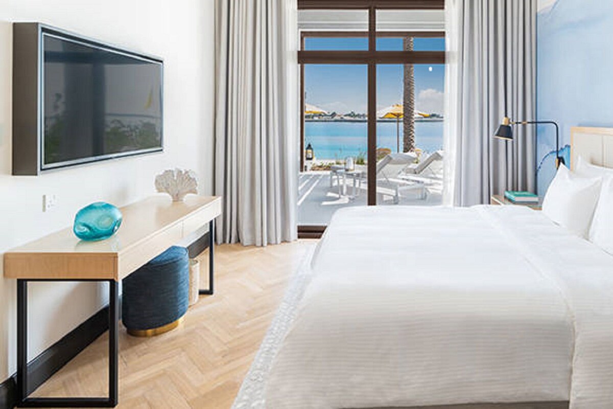Habitación de hotel con cama, escritorio, televisor, vista al mar y balcón con sillones de descanso.