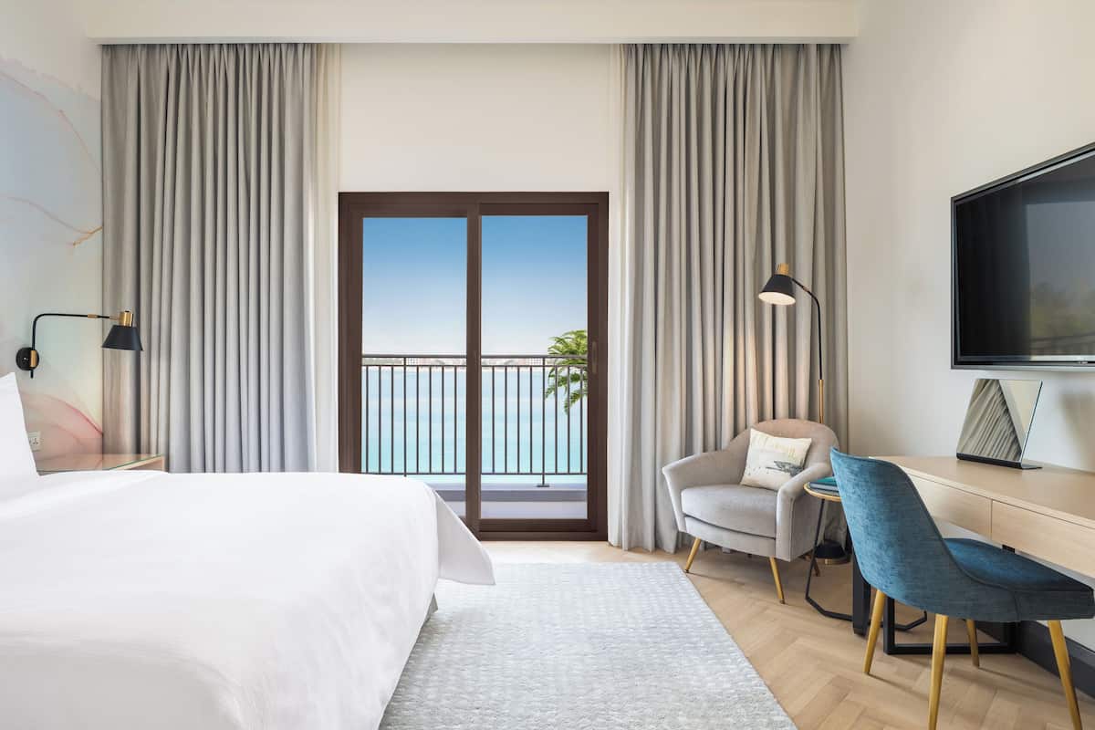 Habitación de hotel con cama, escritorio, silla y un gran ventanal con vistas a un balcón.