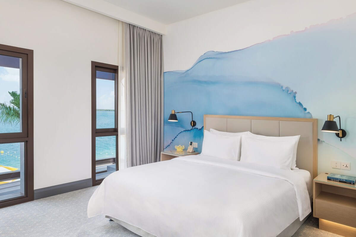 Habitación de hotel con una cama grande, una mesita de noche con lámpara, un mural en la pared y vista al mar a través de amplias ventanas.