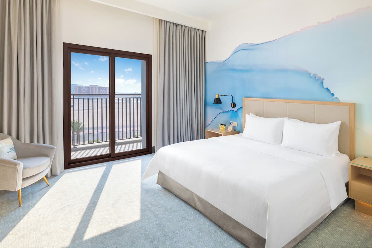 Habitación de hotel con una cama grande, una silla, una mesita de noche y vistas a un balcón.