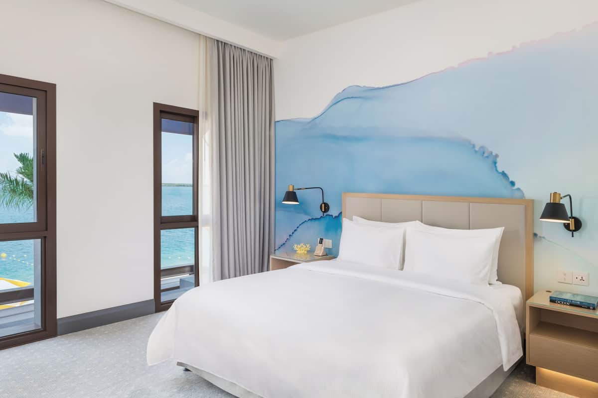 Habitación de hotel con una cama grande, una mesita de noche con lámpara, un mural en la pared y vista al mar a través de amplias ventanas.