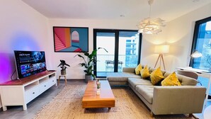 Ein modernes Wohnzimmer mit einem Flachbildfernseher, einem Sofa mit gelben Kissen, einem Couchtisch aus Holz und einem großen Fenster mit Blick auf die Stadt.