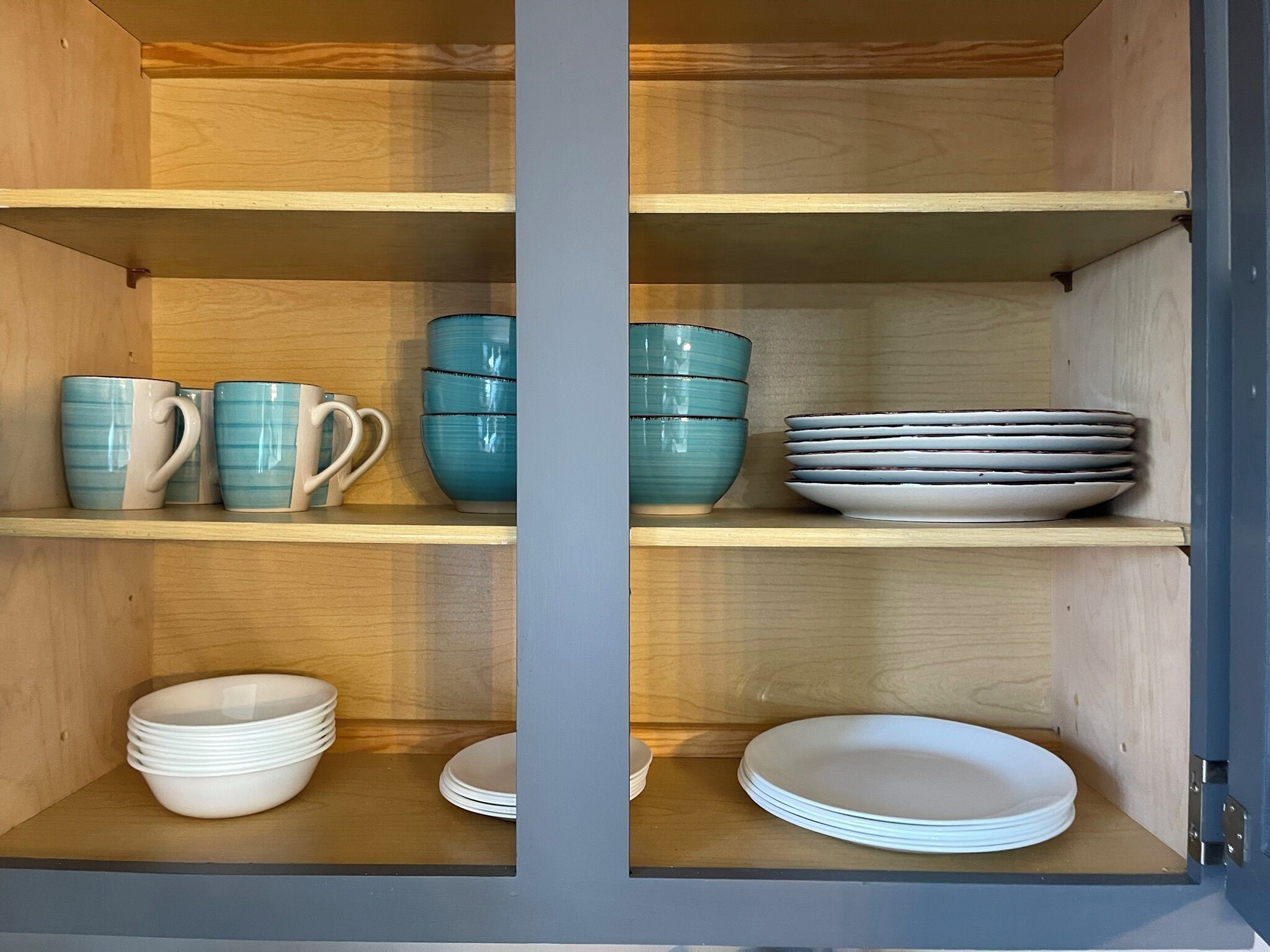 Un mueble con estantes que contiene platos y tazones blancos apilados, y tazas de cerámica azul y blanca.