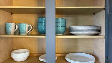 Un mueble con estantes que contiene platos y tazones blancos apilados, y tazas de cerámica azul y blanca.