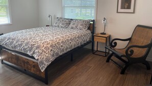 Un dormitorio con cama, dos mesitas de noche, una silla y un cuadro enmarcado en la pared.