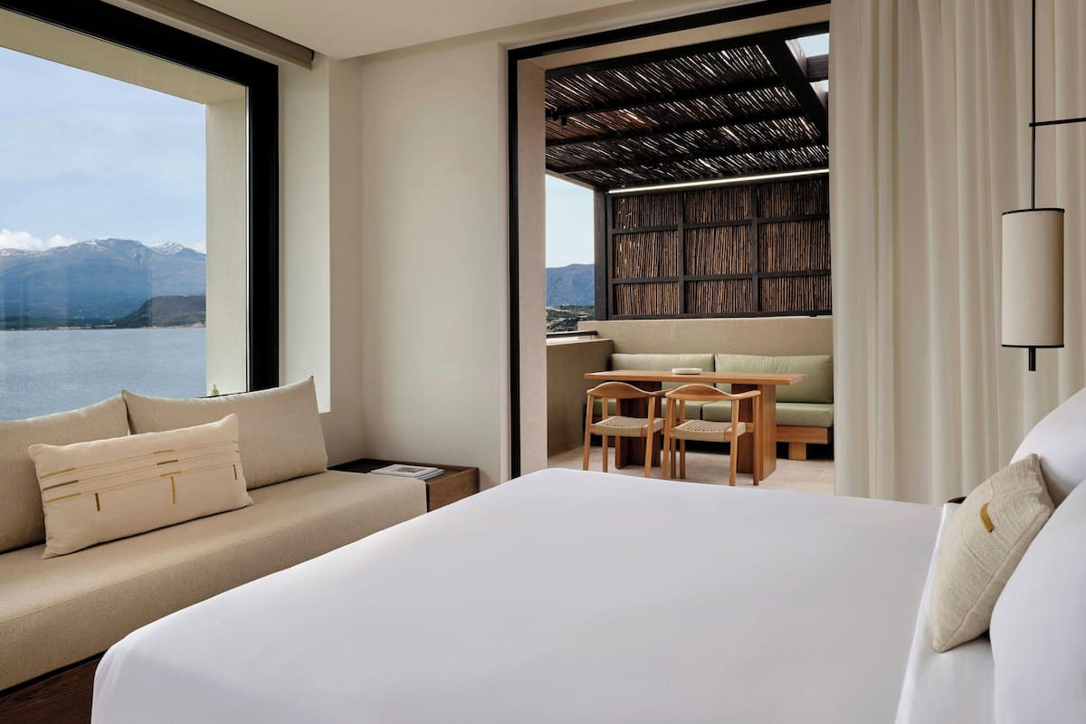 Habitación de hotel con una cama grande, un sofá, un comedor con mesa y sillas, y vistas a las montañas y al agua a través de un ventanal amplio.