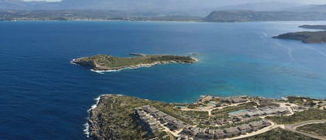 An aerial view of a coastal resort with multiple buildings, a marina, and a mountainous backdrop.