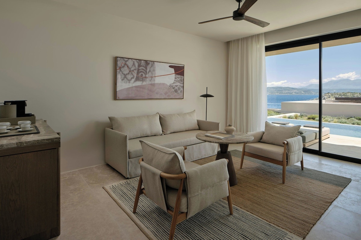 A modern hotel room with a sofa, chairs, a small table, a ceiling fan, and a large window offering a view of the sea.