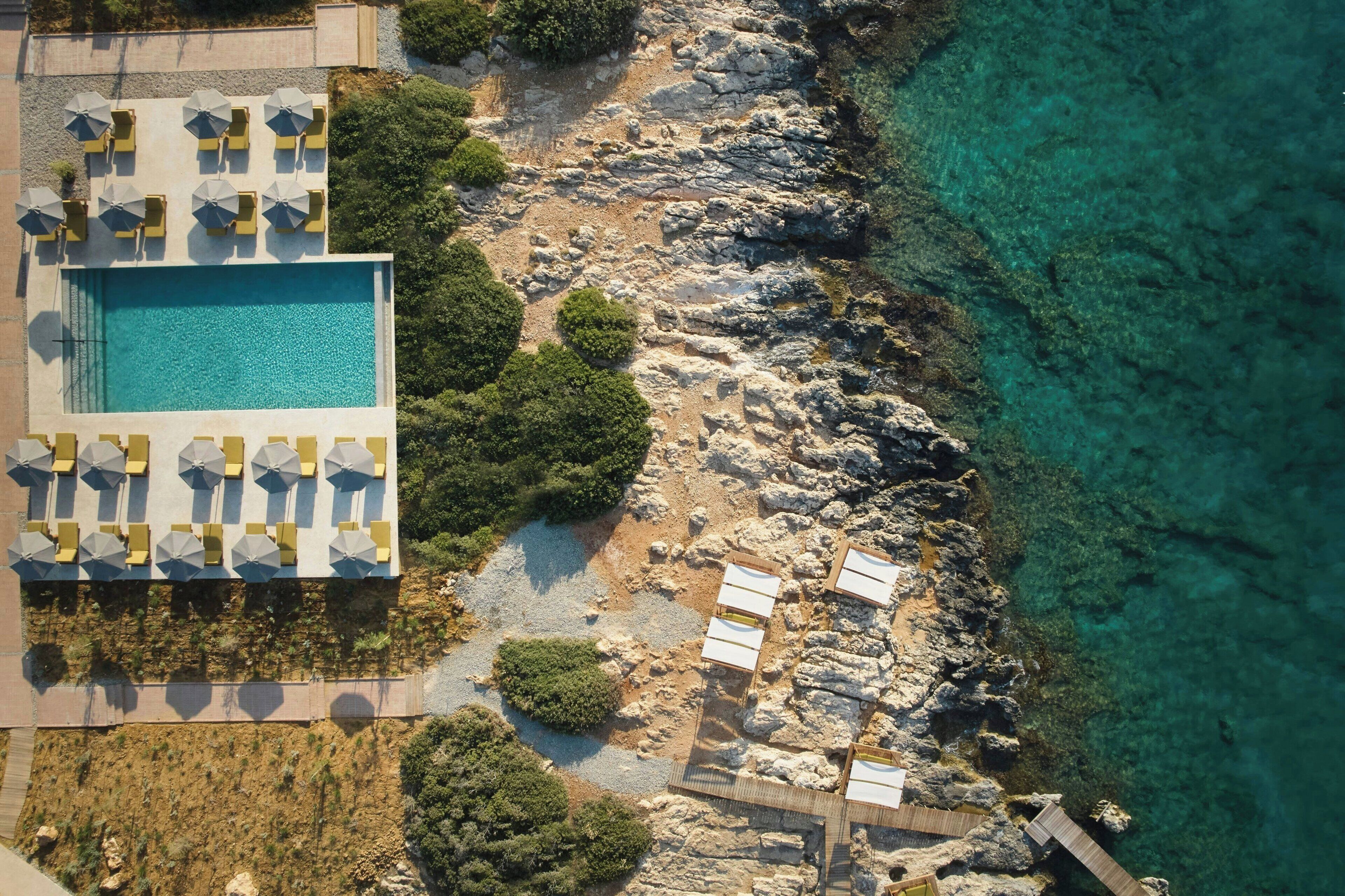 An aerial view of a resort with a swimming pool, sun loungers, and a rocky coastline.