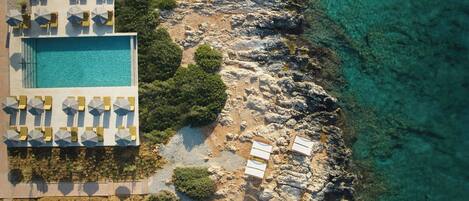 An aerial view of a resort with a swimming pool, sun loungers, and a rocky coastline.
