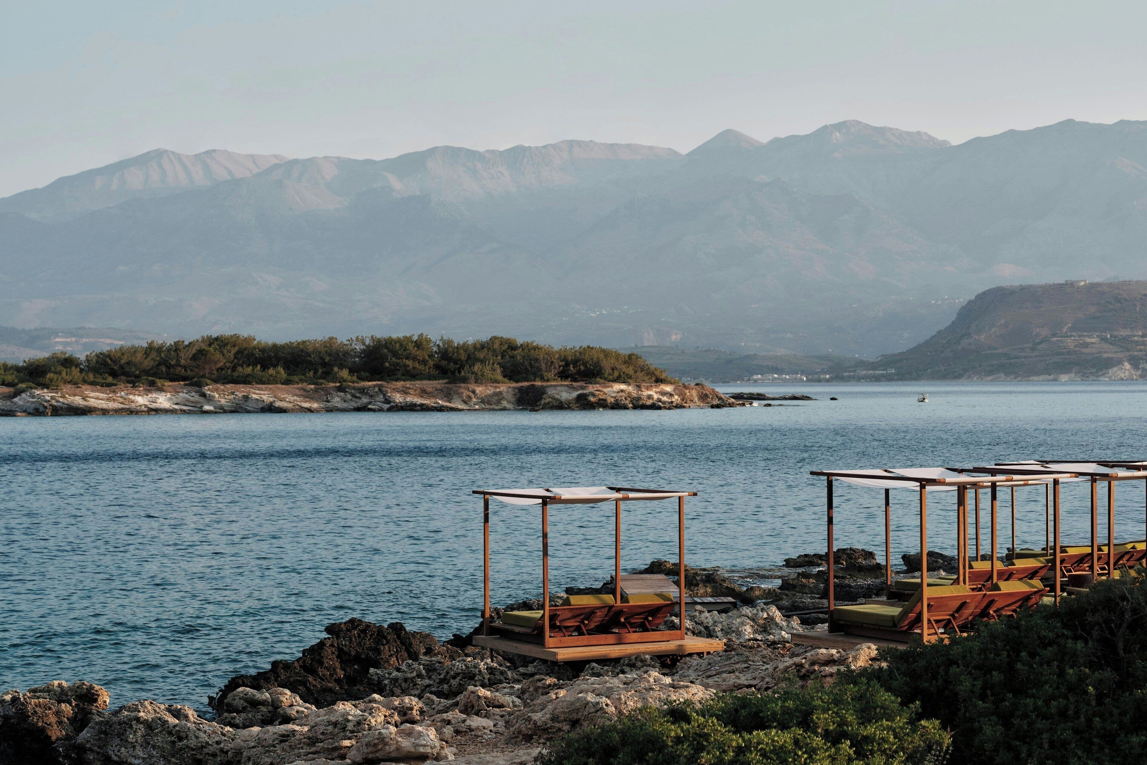 A coastal area with a rocky shoreline, two wooden gazebos with seating, and a view of distant mountains.