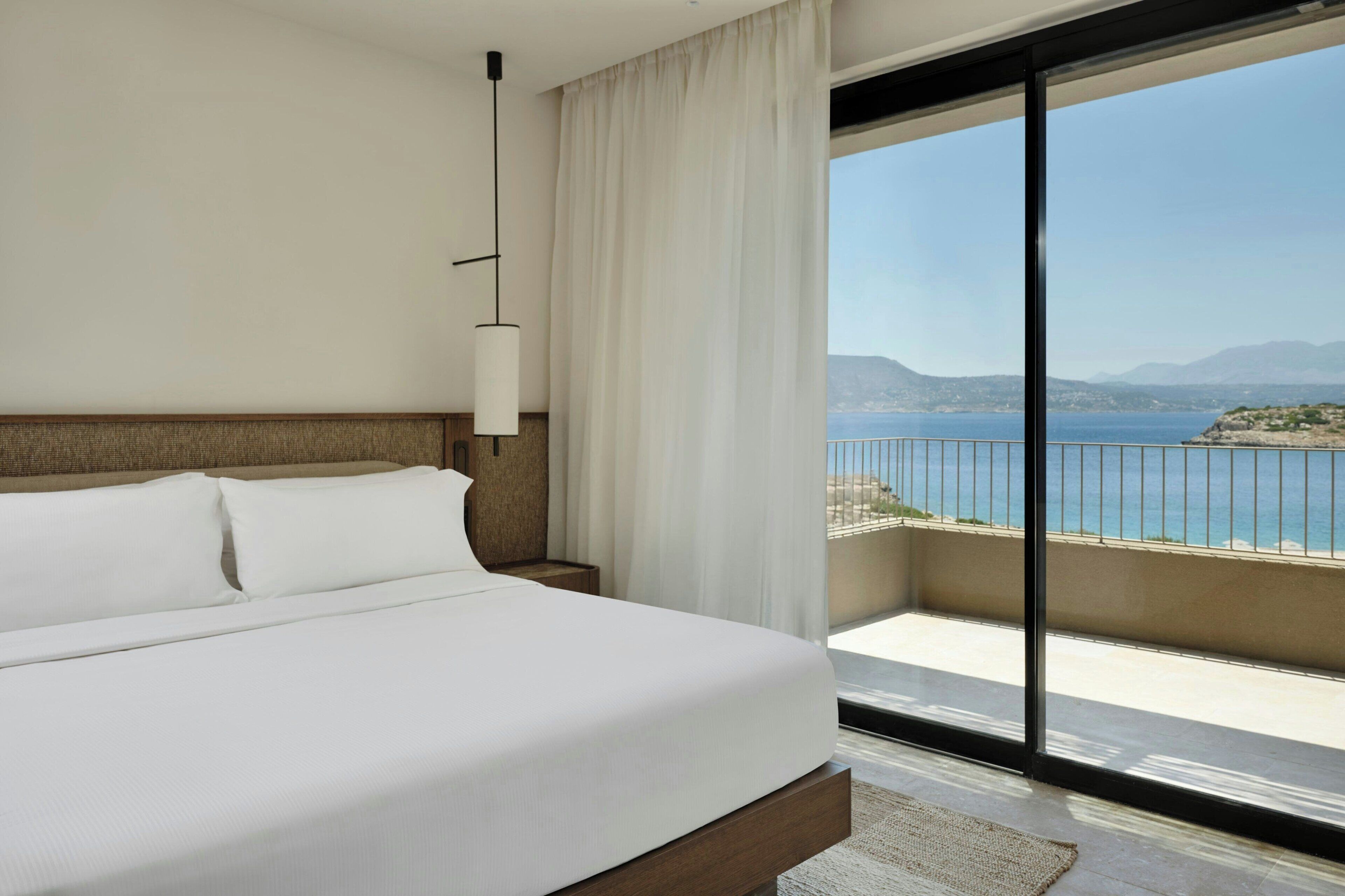 A hotel room with a large bed, a nightstand, a lamp, and a view of the sea through a balcony.