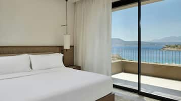 A hotel room with a large bed, a nightstand, a lamp, and a view of the sea through a balcony.