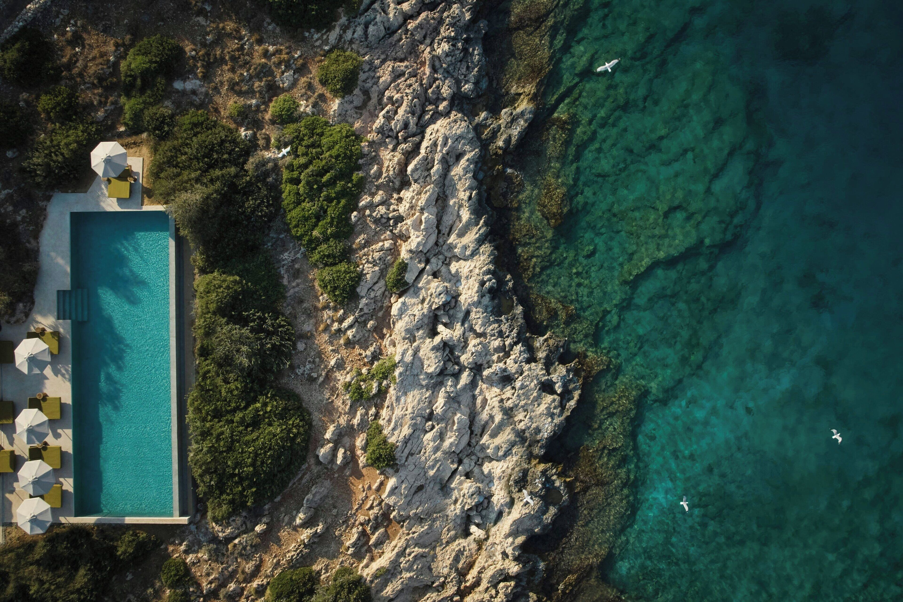 An aerial view of a coastal property with a rectangular swimming pool, surrounded by lush greenery and rocky terrain.