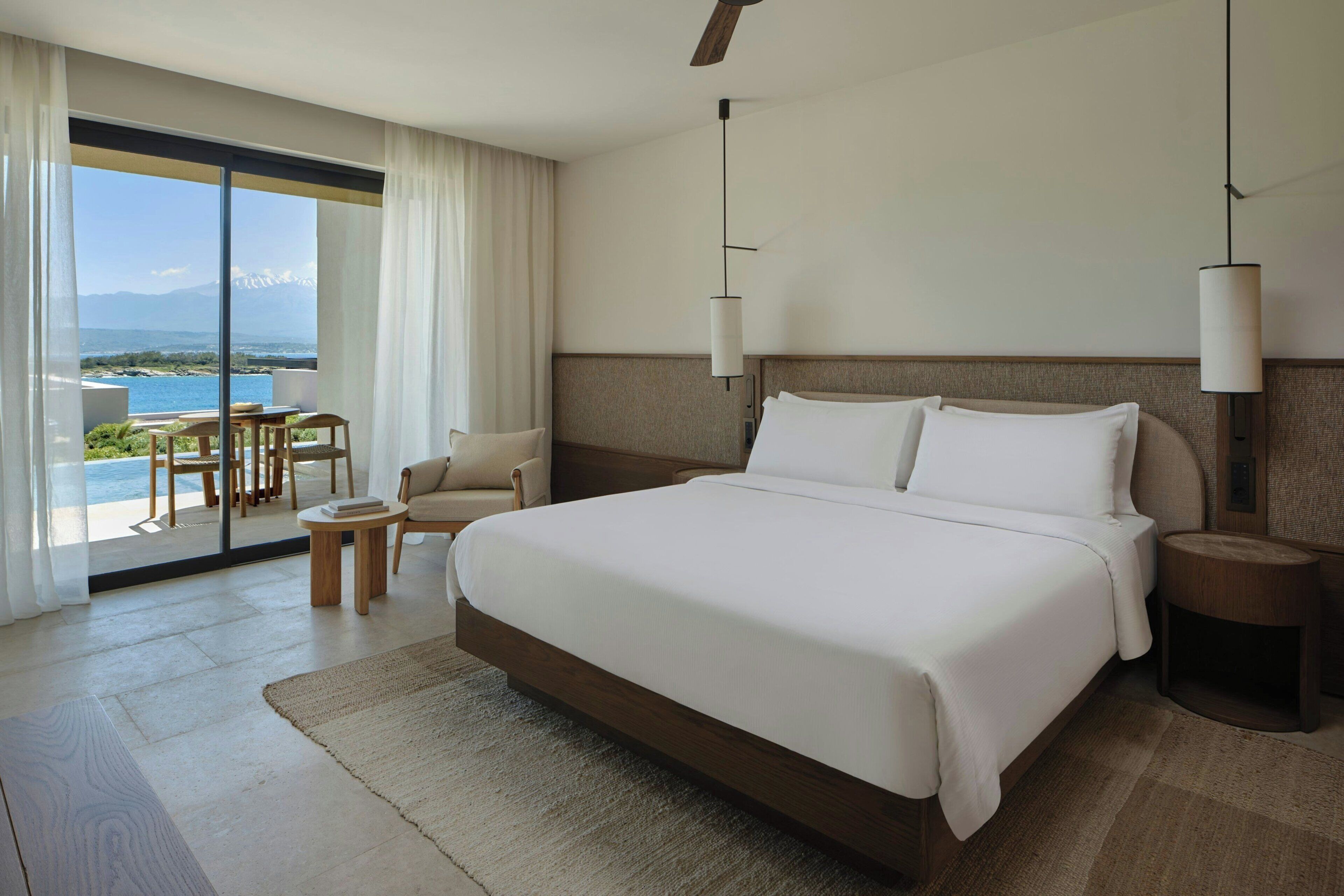 Ein modernes Hotelzimmer mit einem großen Bett, einem kleinen runden Tisch, einem Sessel und Blick auf einen Pool und Berge.