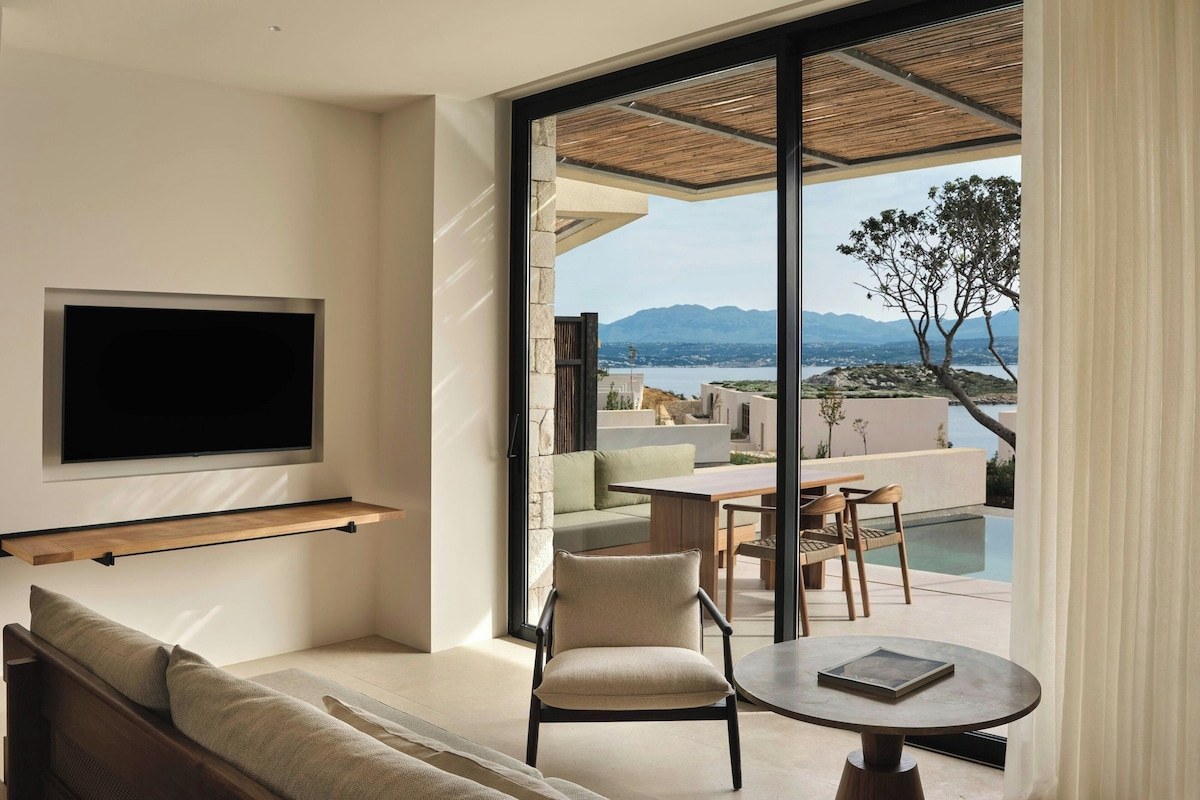 A modern living room with a flat-screen TV, a sofa, a chair, and a table. There is a large window with a view of a pool and mountains.